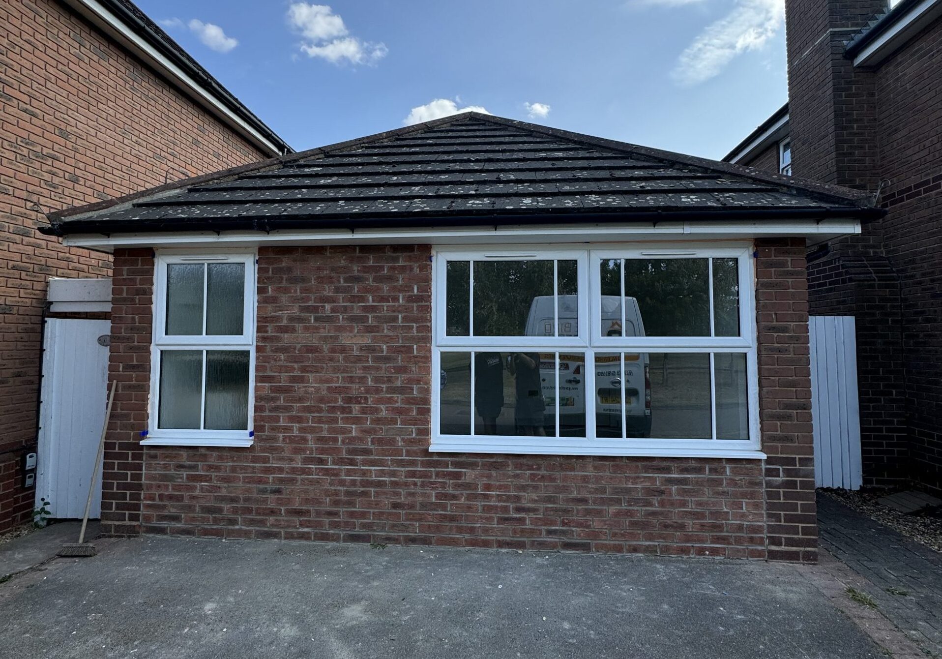 A small brick building with a dark-tiled roof, white-framed windows, and a concrete drive in front. A broom leans against the left wall, and the reflection reveals a white van—ideal for kitchen and bathroom showroom installation and design supply.