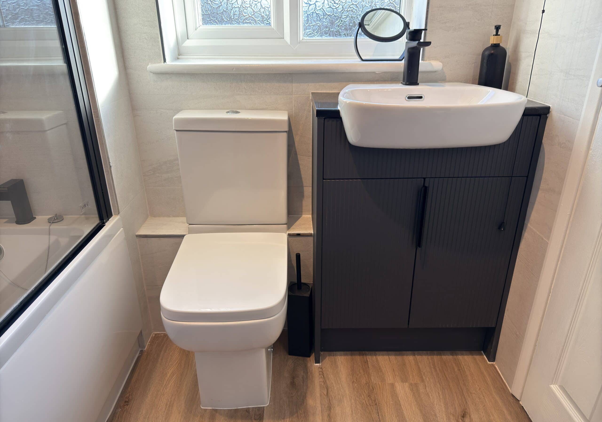 Modern bathroom with a white toilet next to a black cupboard sink, black tap, soap dispenser, and small mirror. Wood-style floor and frosted window highlight sleek design—perfect inspiration from a kitchen and bathroom showroom.