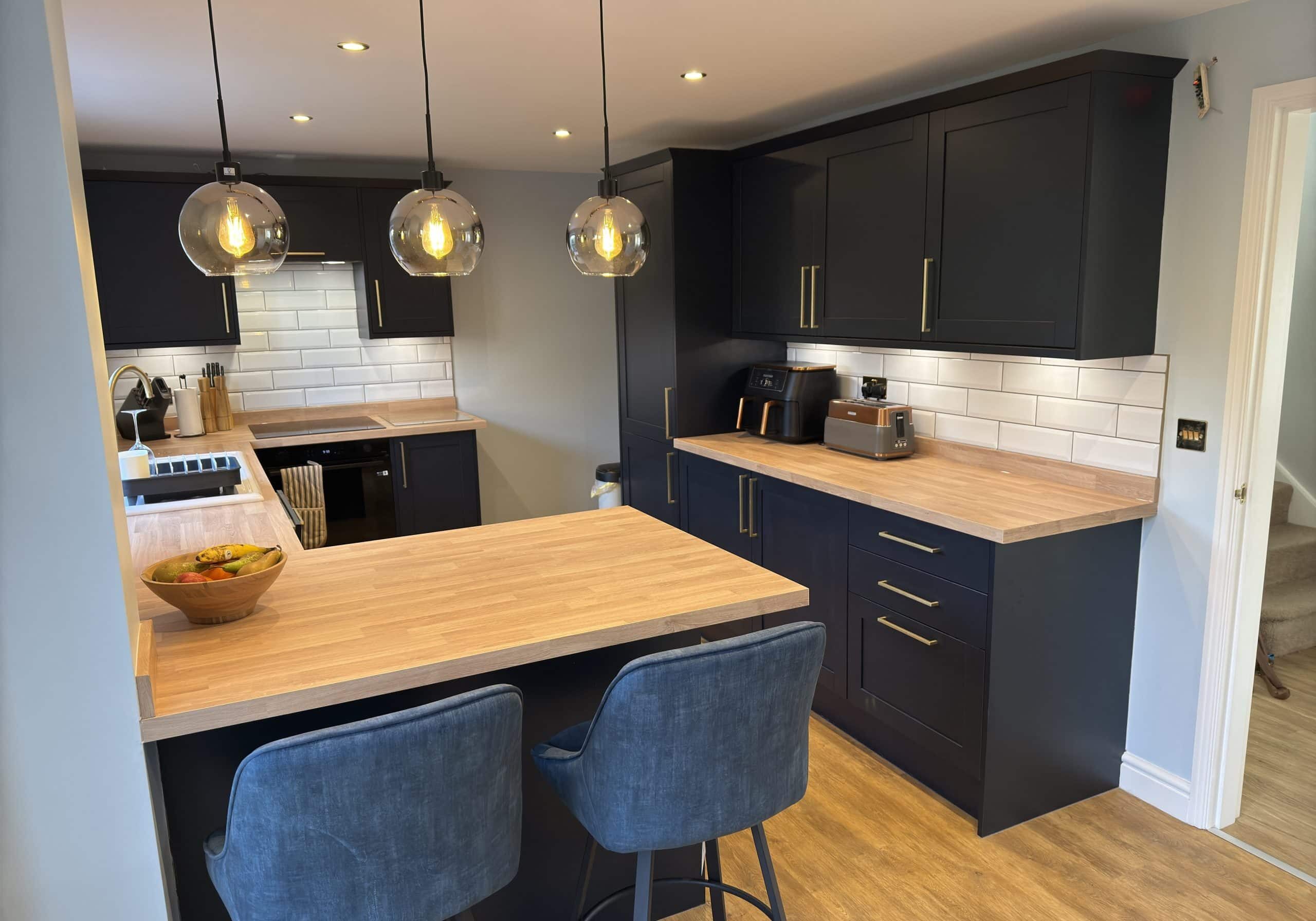 Modern kitchen with navy cupboards, wooden worktops, white tiled splashback, and hanging pendant lights. Two blue barstools sit at the counter, highlighting stylish design found in top kitchen and bathroom showroom installations.