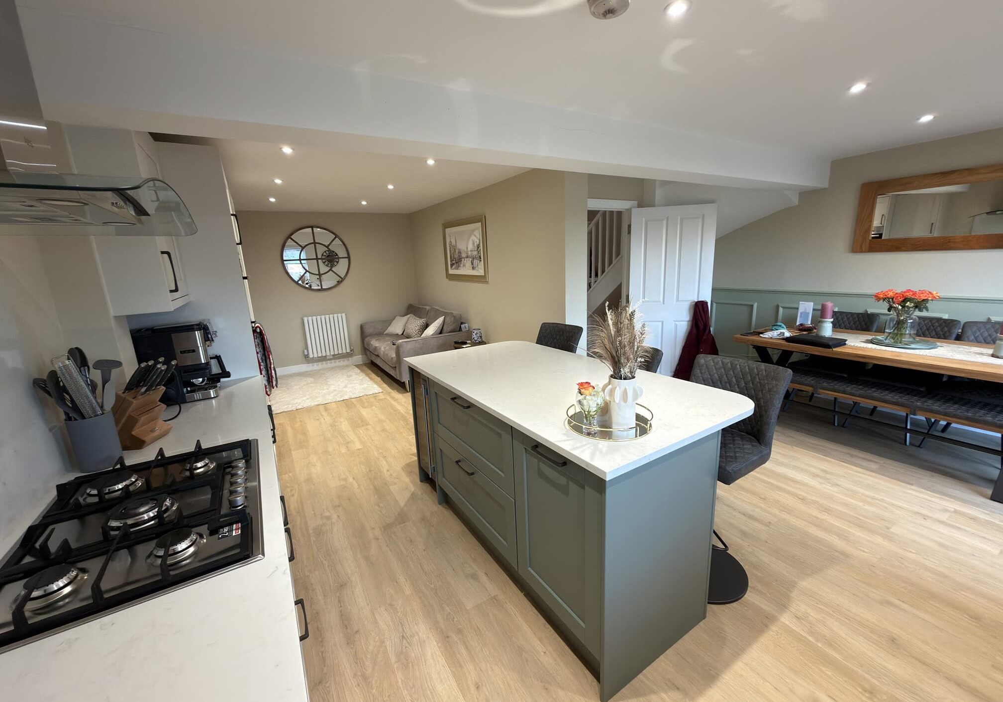 Open-plan kitchen and living area with a central island, gas hob, grey cupboards, wooden floors, dining table with bench seating, a sofa, large clock on the wall, and neutral décor.