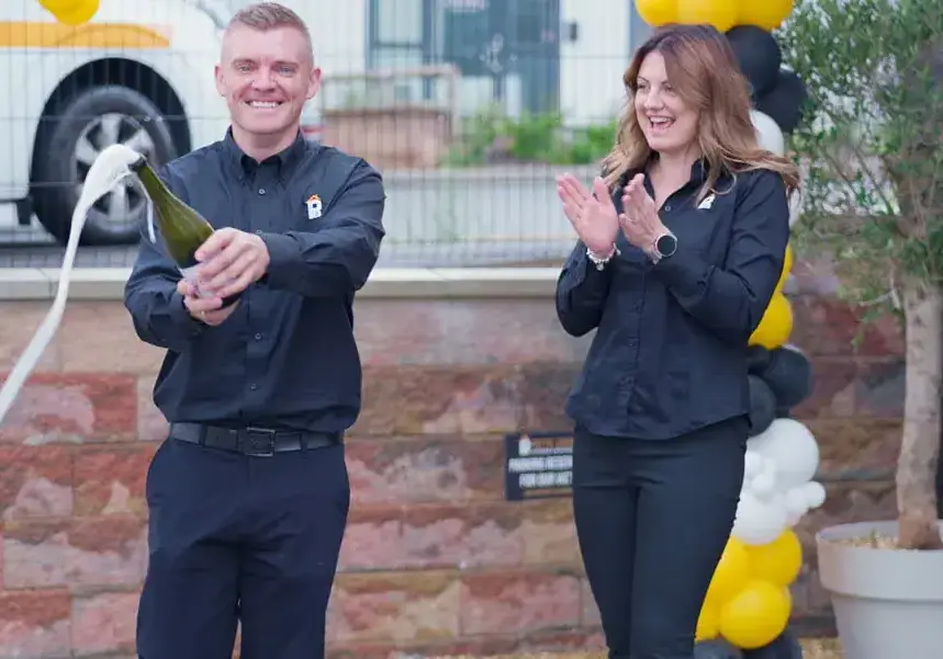 A smiling man in a black shirt pops a bottle of champagne as foam sprays out, while a woman in a black outfit claps. They celebrate outdoors near a stone wall and black and yellow balloons after a successful kitchen and bathroom showroom installation.