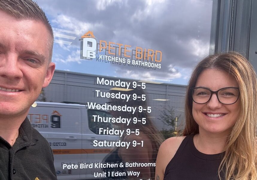 Two people are smiling in front of a glass door with the Pete Bird Kitchens & Bathrooms logo and business hours displayed, reflecting the sky and nearby building—a welcoming spot for kitchen and bathroom showroom design inspiration.