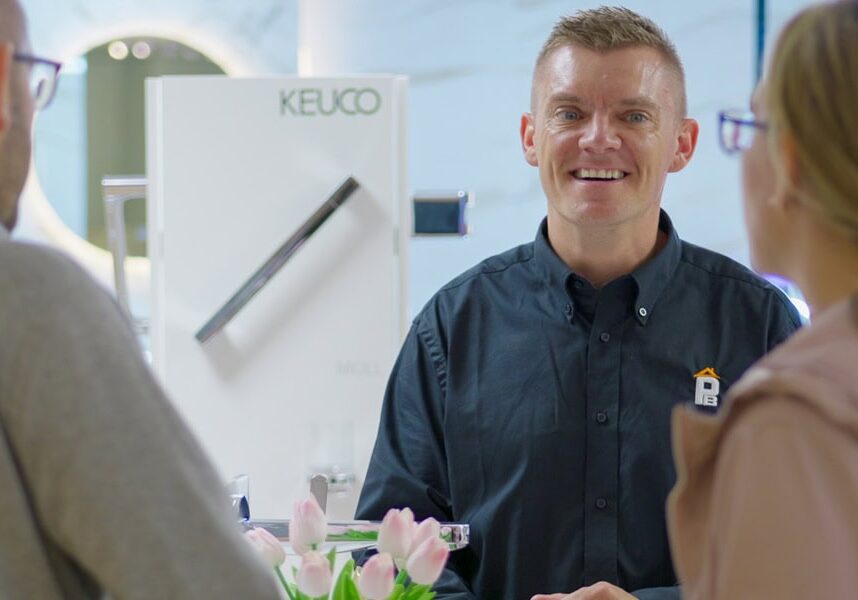 A smiling sales assistant in a black shirt talks to two customers at a counter.