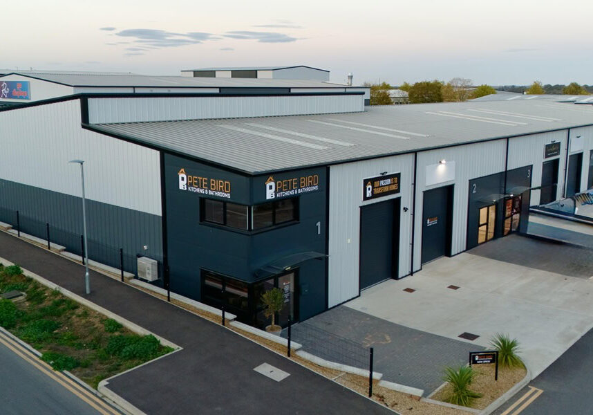 A modern industrial building with grey and white cladding, large windows, and Pete Bird Construction signs. Several parked vehicles and a paved pavement surround the structure. The sky is partly cloudy.