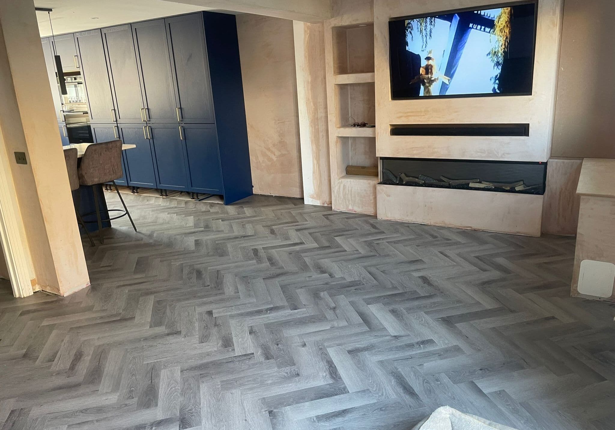 A modern living room with light grey herringbone flooring, a wall-mounted TV showing a person, a built-in fireplace, light walls, and blue kitchen cupboards—perfect inspiration for any kitchen and bathroom showroom design.