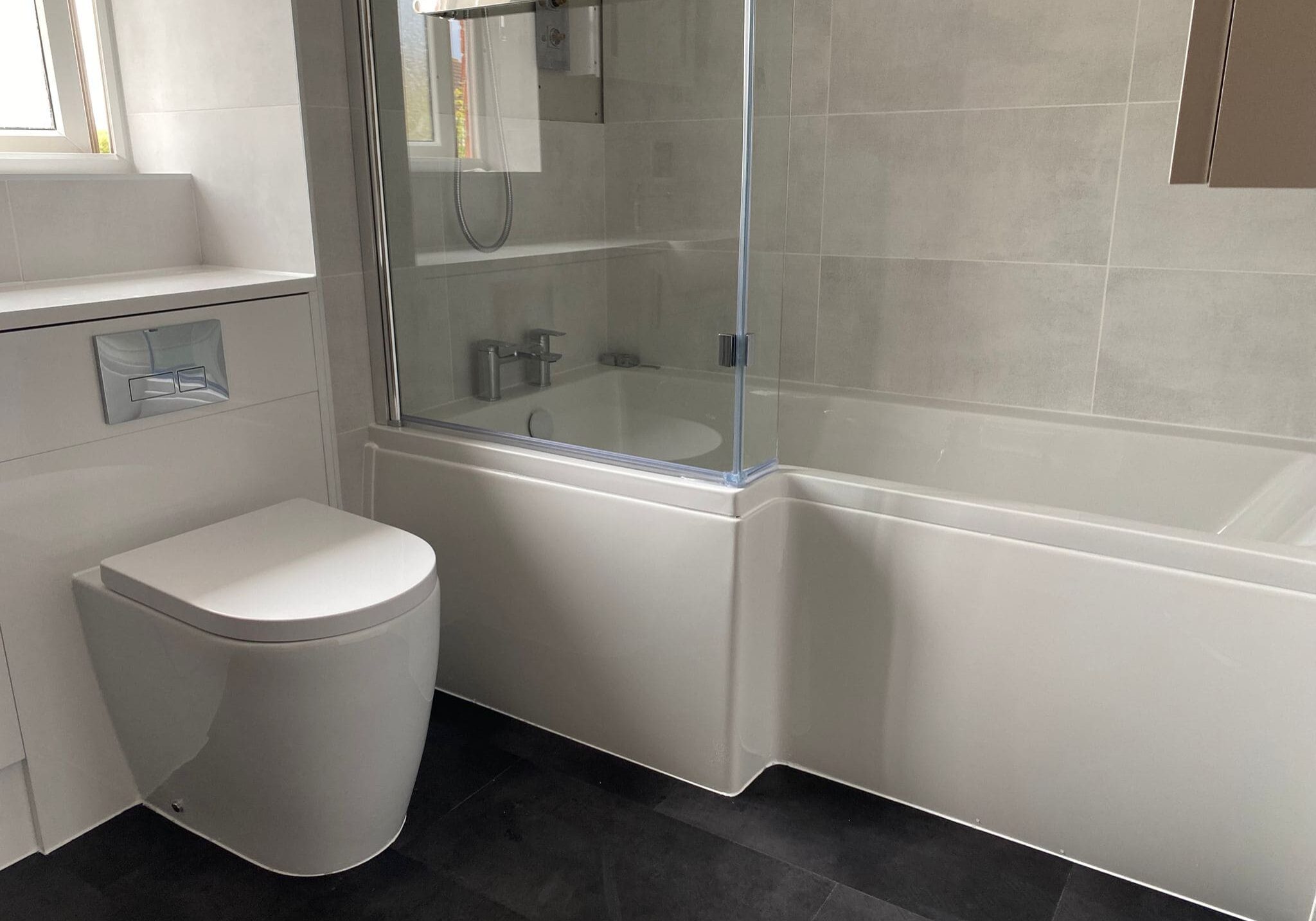 A modern bathroom designed with a white toilet, a white bath with a glass shower screen, silver fittings, light grey wall tiles, and a black floor—showcasing stylish installation from our kitchen and bathroom showroom. Natural light flows in through the window.